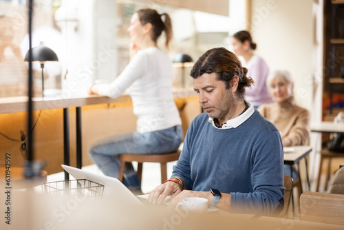 Focused adult man sitting at table with cup of coffee and laptop, browsing websites on internet