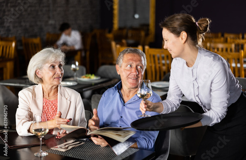 Amiable female owner of restaurant bringing glasses of wine to positive senior couple, sitting at table and choosing dishes from menu card