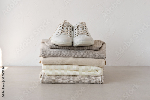 Minimalist pastel neutral beige autumn knitware clothing stack, sneakers on table, on white wall background. Aesthetic fashion woman fall wardrobe, business brand, blog template