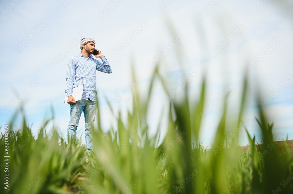 Portrait of happy young Indian man farmer standing in agriculture land surround by crop holding smartphone talking to someone, smiling village male with mobile phone in field, technology concept Portrait of happy young Indian man farmer standing in agriculture land surround by crop holding smartphone talking to someone, smiling village male with mobile phone in field, technology concept