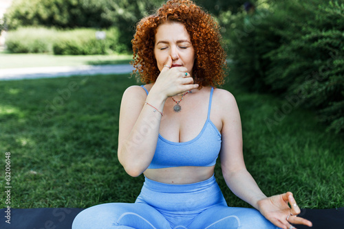 Pranayama and yoga practice outdoor, stress relief techniques. Redhead girl with curly hair sitting with crossed legs on mat in park with closed eyes, closing one nostril with thumb to reduce anxiety