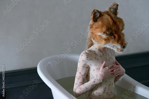 Young woman in wet bodysuit and fox mask on her head sitting straight in tub and covering breast with her hands as if protecting from outside world  