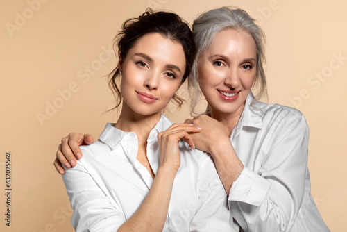 Charming beautiful mother and daughter in white casual shirt studio face forward portrait. Affectionate senior woman hugging adorable young girl shoulder from back. Adult child touching mom hand