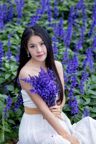Asian woman in white dress sitting amidst in flowers field. Beautiful girl holding bouquet flowers in hands. Happy woman enjoying in purple flower field and nature sea of fog. Embrace nature wind.