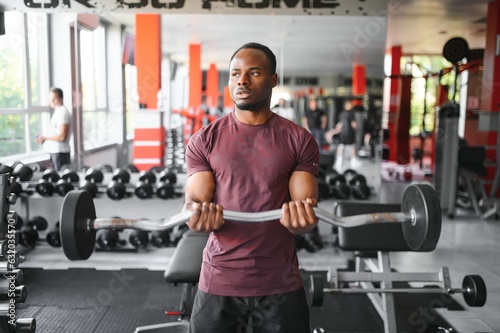 Hot african american young man bodybuilder lifting barbell at gym, working on his arms, looking at copy space. Black muscular shirtless guy having biceps workout session. Healthy lifestyle concept.