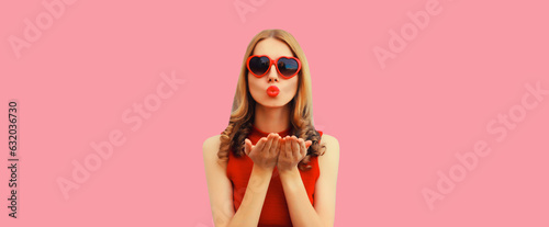 Summer portrait of beautiful young woman model blowing her lips sending sweet air kiss wearing red heart shaped sunglasses on pink background