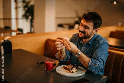 A man in a casual outfit sitting in the cafe and eating a burger.