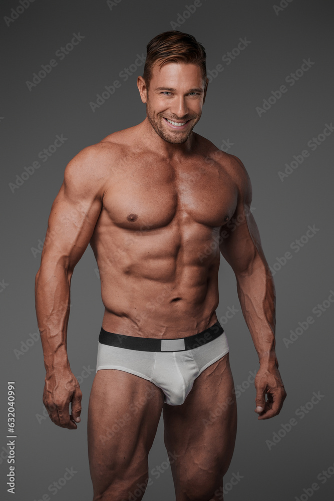 A muscular and charismatic male model posing confidently in underwear, flashing a bright smile against a grey backdrop