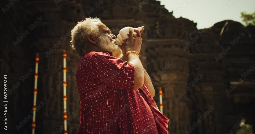 Authentic Shot of An Indian Senior Monk Blowing a Conch Shell in an Ancient Stone Temple. Elderly Man Performing Sacred Ritual on a Worship Day. Hinduism, South Asian Religion and Cultural Footage