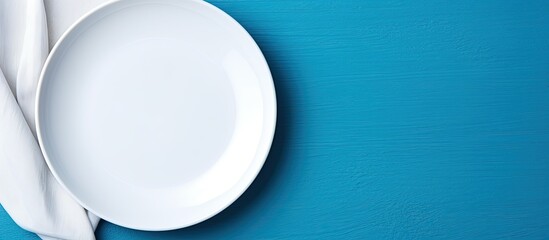 This image is of an empty white plate and a cotton napkin on a blue background. It is a food background suitable for a menu or recipe book. The table setting is shown from a top-down view, and is