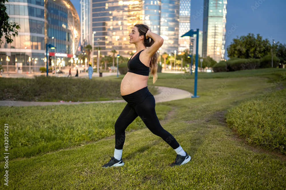 Side view of pregnant woman in sportswear doing lunges in a park at evening.