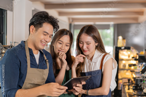 Three barista entrepreneur team meeting at coffee shop. Young smiling Asian male and female cafe employees having discussion on work and menu with tablet computer