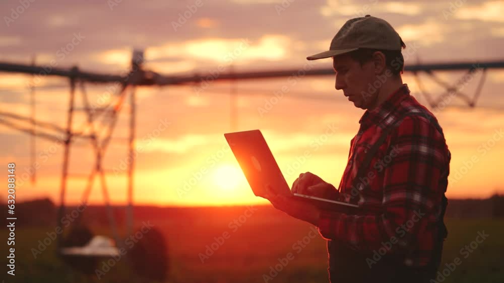 silhouette agriculture. a male farmer works on a laptop in a field with green corn sprouts. corn is watered by irrigation machine. irrigation agriculture sunlight business concept