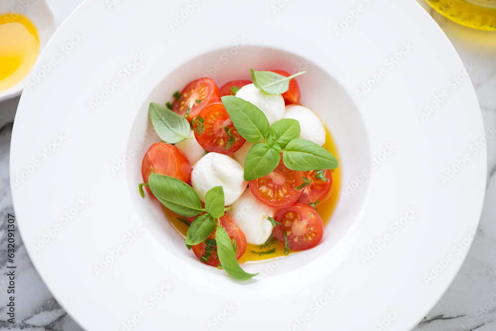 Middle close-up of caprese salad with baby mozzarella, cherry tomatoes, olive oil and green basil served in a white plate Middle close-up of caprese salad with baby mozzarella, cherry tomatoes, olive oil and green basil served in a white plate