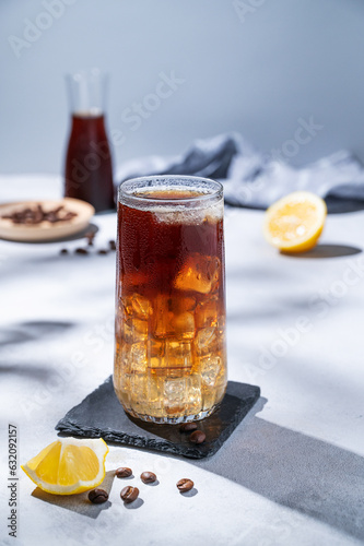 Coffee cold brew with tonic in a tall glass with ice and lemon on a light blue background with coffee beans   bottle  fruits and shadows. Trendy summer craft refreshing drink.