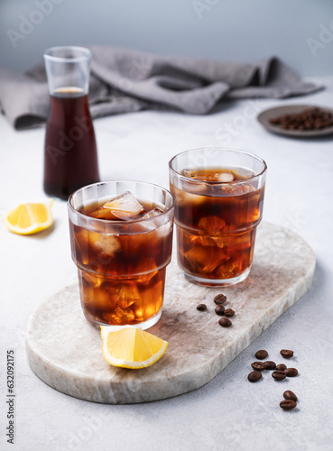 Cold brew coffee in a two glasses with lemon and ice on a marble board on a light background with coffee beans, bottle and napkin. Concept summer craft refreshing homemade drink