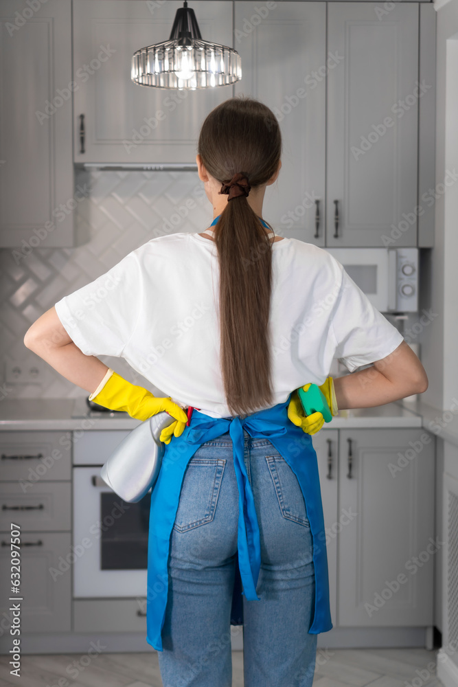 Rear view of a young woman in an apron, protective gloves, holding a spray and sponge while cleaning in the kitchen. Concept of housework, cleaning company. Vertical photo Rear view of a young woman in an apron, protective gloves, holding a spray and sponge while cleaning in the kitchen. Concept of housework, cleaning company. Vertical photo