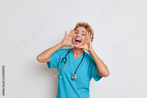 Happy female doctor posing in white studio with hands at her mouth like loud-hailer and smiling, good doctor concept, copy space