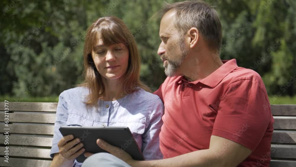 A middle-aged Caucasian couple looks at a tablet and smiles as they sit on a bench in a park on a sunny day
