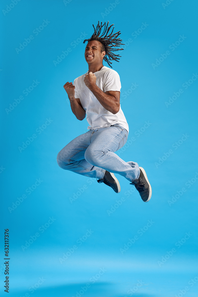 Fototapeta premium Reaching success. Full-length image of young man in casual clothes jumping with happy emotions against blue studio background. Concept of youth, human emotions, lifestyle, fashion, ad