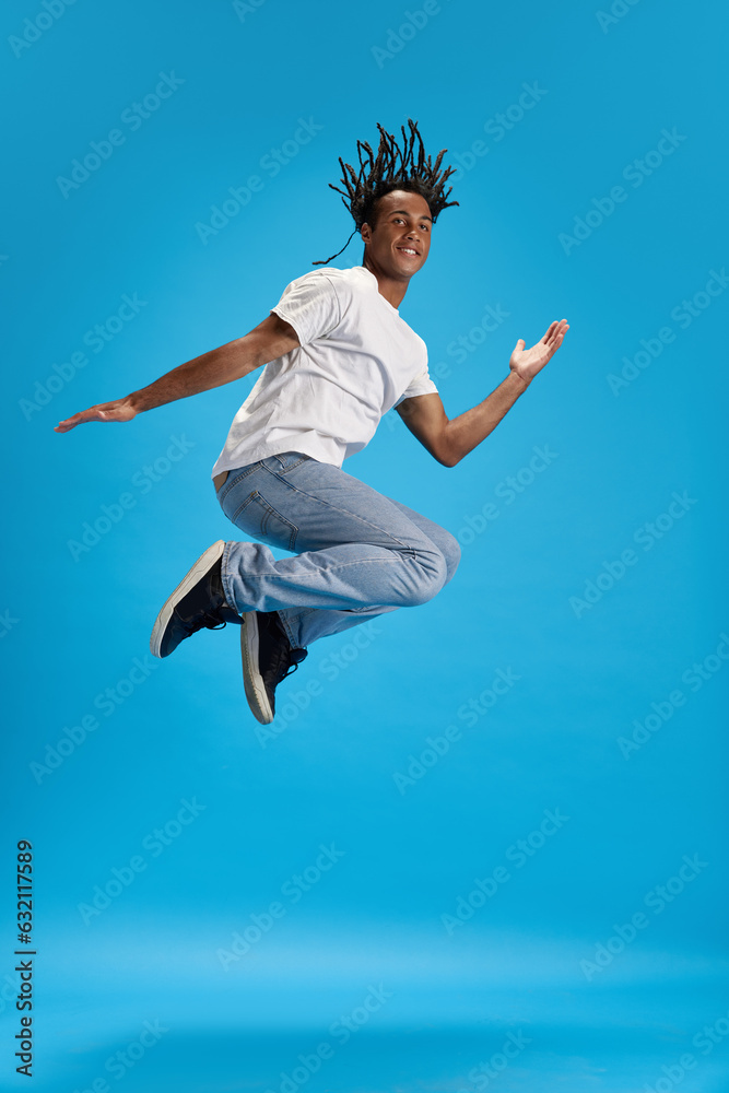 Full-length image of young african man with dreads in casual clothes jumping against blue studio background. Feeling positive. Concept of youth, human emotions, lifestyle, fashion, ad
