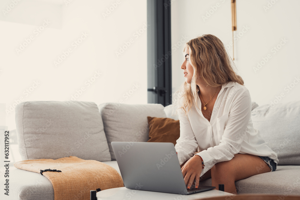Portrait of one young attractive blonde woman using laptop pc computer on couch relaxing surfing the net at home looking at the window waiting package Portrait of one young attractive blonde woman using laptop pc computer on couch relaxing surfing the net at home looking at the window waiting package