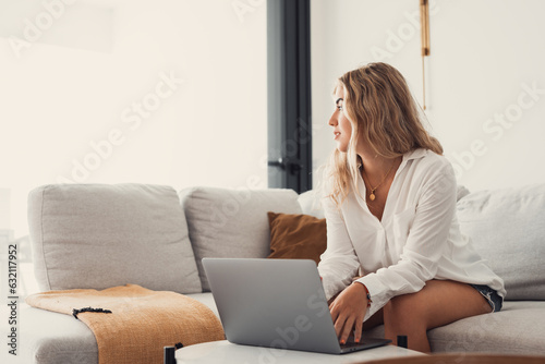 Portrait of one young attractive blonde woman using laptop pc computer on couch relaxing surfing the net at home looking at the window waiting package