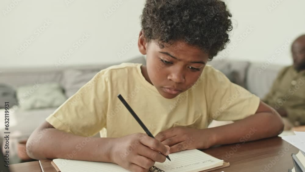 Chest up of African American preteen boy writing in copybook while doing school homework sitting at desk in modern living room Chest up of African American preteen boy writing in copybook while doing school homework sitting at desk in modern living room