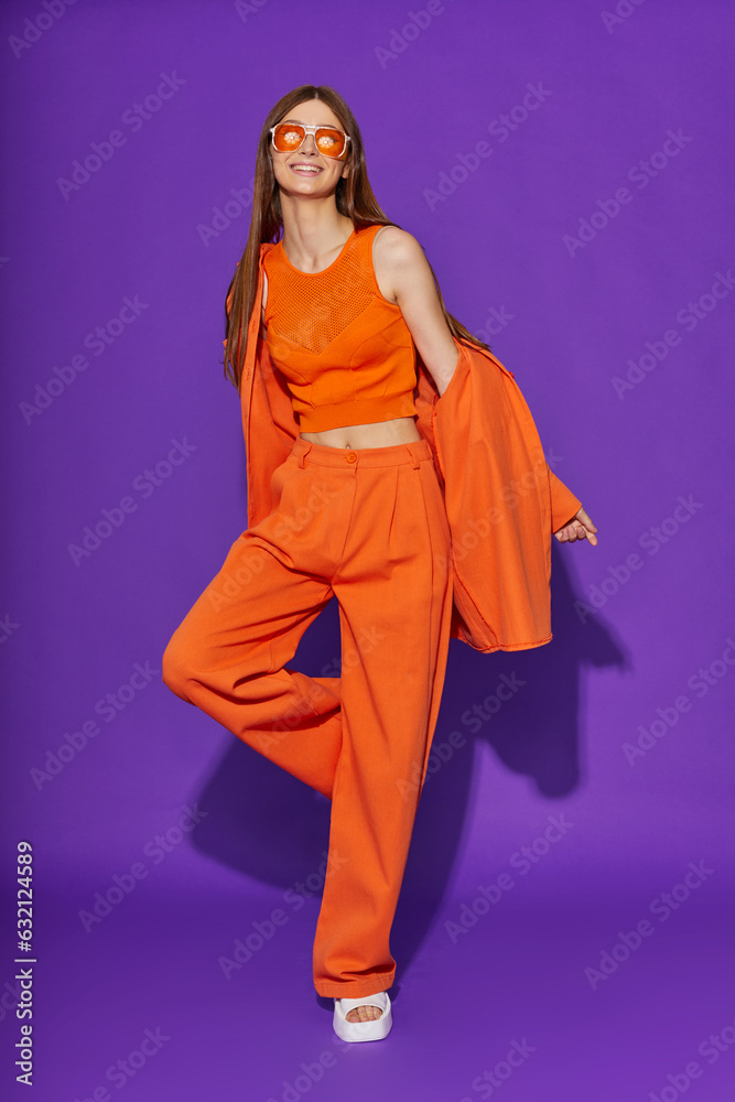 Young fashion woman in orange pants orange top orange shirt on violet background. Platform slides sandals, orange sunglasses.