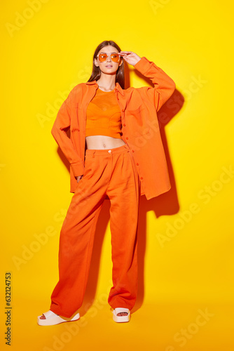 Young fashion woman in orange pants orange top orange shirt on yellow background. Platform slides sandals, orange sunglasses.