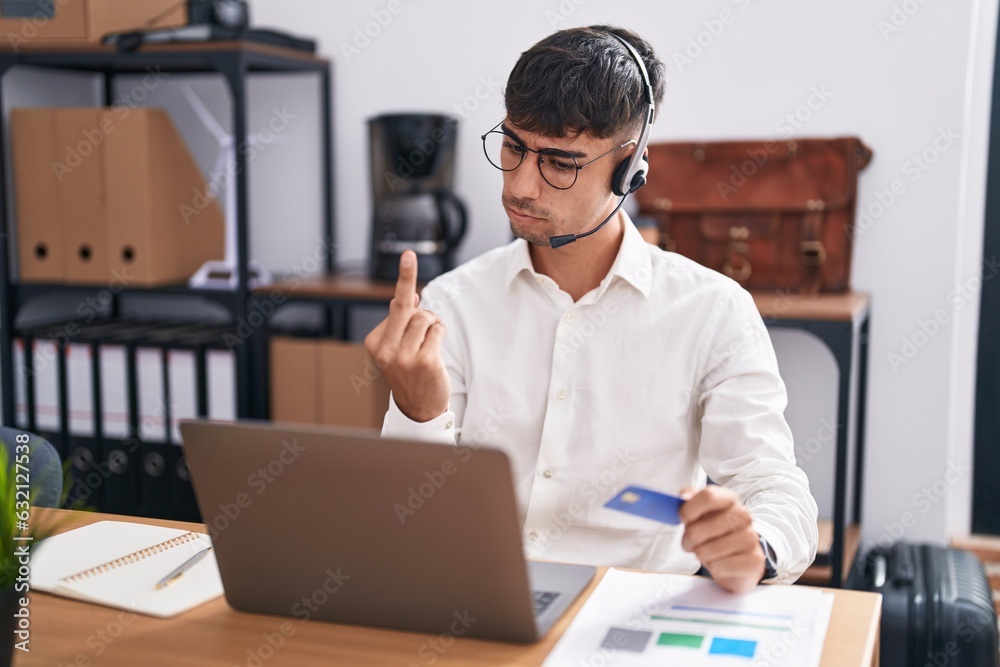 Young hispanic man working using computer laptop holding credit card showing middle finger, impolite and rude fuck off expression Young hispanic man working using computer laptop holding credit card showing middle finger, impolite and rude fuck off expression