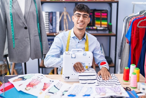 Young hispanic man tailor smiling confident using sewing machine at tailor shop
