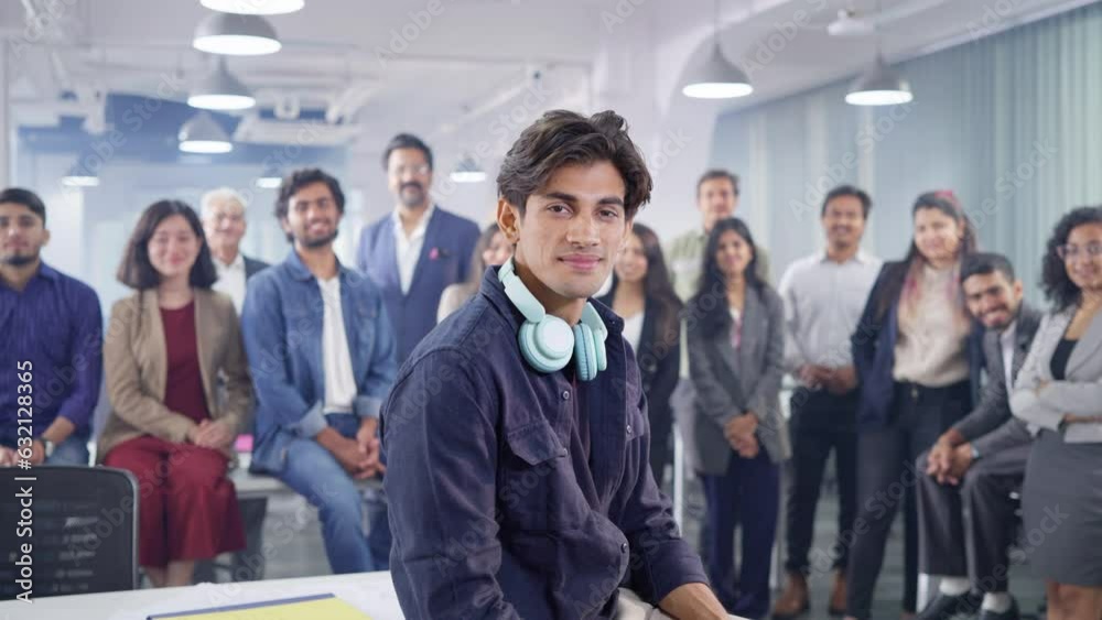Indian Asian happy modern male stylish hipster or young man wearing headphones around neck looking at camera smiling with team of business executives standing in the back in corporate start up office. Indian Asian happy modern male stylish hipster or young man wearing headphones around neck looking at camera smiling with team of business executives standing in the back in corporate start up office.
