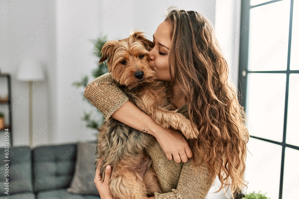 Young beautiful hispanic woman kissing and hugging dog standing at home Young beautiful hispanic woman kissing and hugging dog standing at home