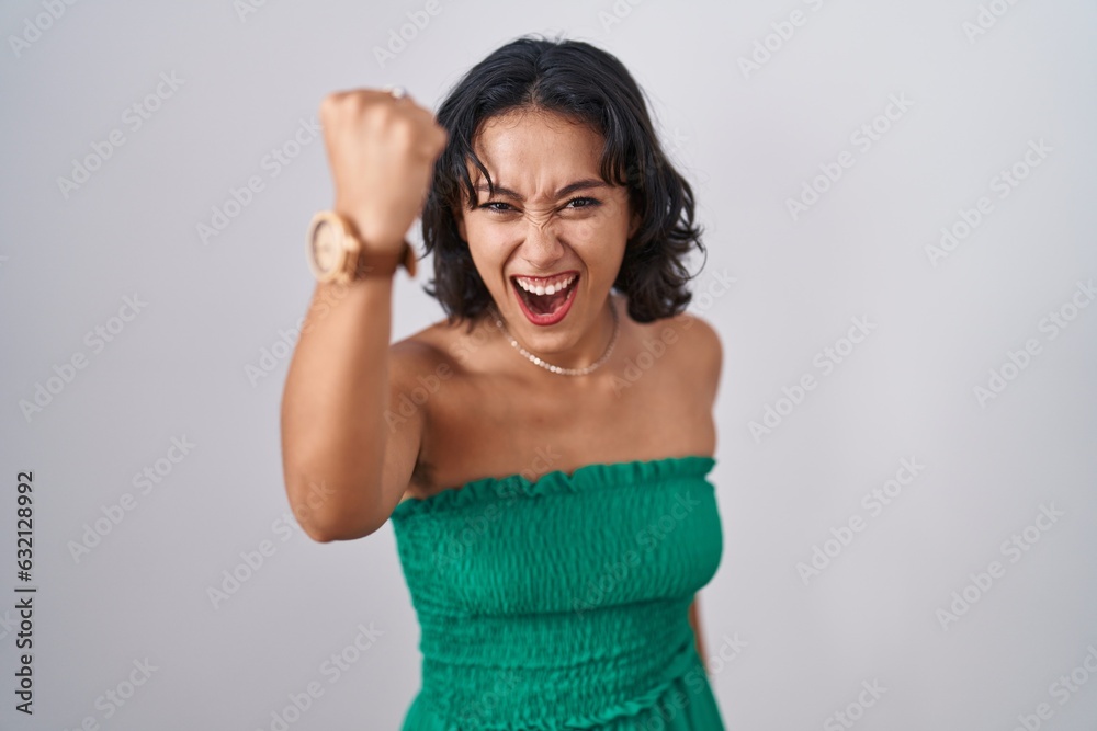 Young hispanic woman standing over isolated background angry and mad raising fist frustrated and furious while shouting with anger. rage and aggressive concept.