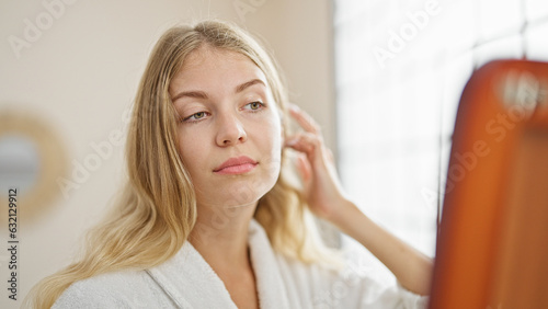 Young blonde woman combing hair looking on mirror at bedroom