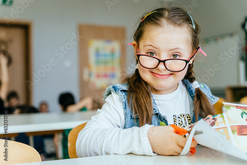 Smiling junior cauasian school girl with down syndrome sitting at desk in classroom, cutting paper with scissors, posing and looking at camera. photo
