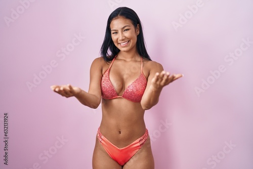 Hispanic woman wearing bikini smiling cheerful offering hands giving assistance and acceptance.