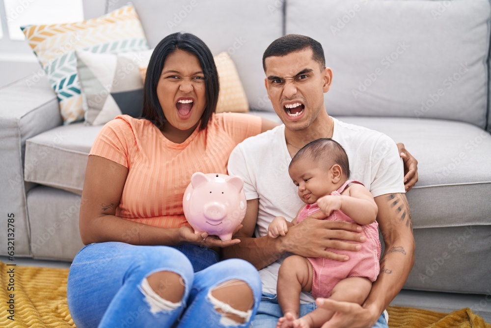 Young hispanic couple with baby holding piggy bank angry and mad screaming frustrated and furious, shouting with anger. rage and aggressive concept. Young hispanic couple with baby holding piggy bank angry and mad screaming frustrated and furious, shouting with anger. rage and aggressive concept.