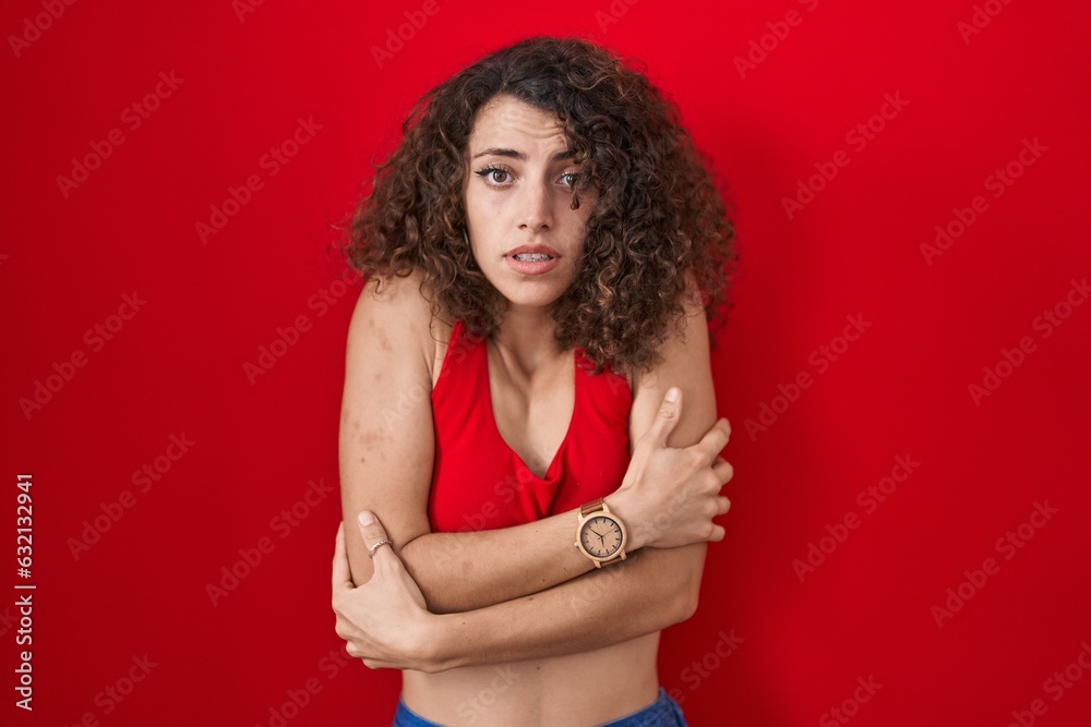 Hispanic woman with curly hair standing over red background shaking and freezing for winter cold with sad and shock expression on face Hispanic woman with curly hair standing over red background shaking and freezing for winter cold with sad and shock expression on face