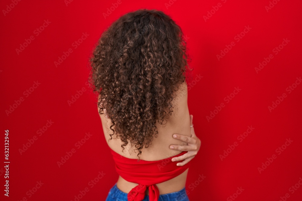 Hispanic woman with curly hair standing over red background hugging oneself happy and positive from backwards. self love and self care Hispanic woman with curly hair standing over red background hugging oneself happy and positive from backwards. self love and self care