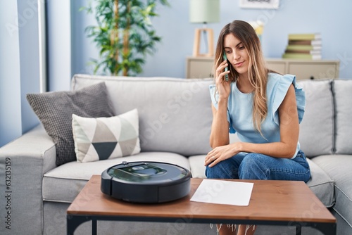 Obraz na plátně Young hispanic woman sitting at home by vacuum robot speaking on the phone think