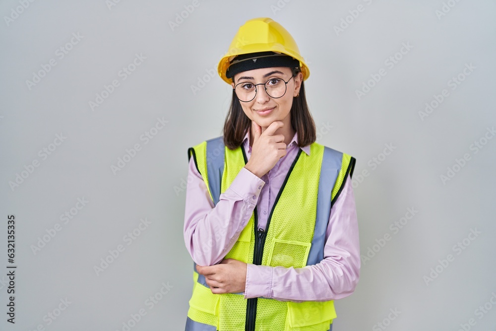 Hispanic girl wearing builder uniform and hardhat looking confident at the camera smiling with crossed arms and hand raised on chin. thinking positive. Hispanic girl wearing builder uniform and hardhat looking confident at the camera smiling with crossed arms and hand raised on chin. thinking positive.