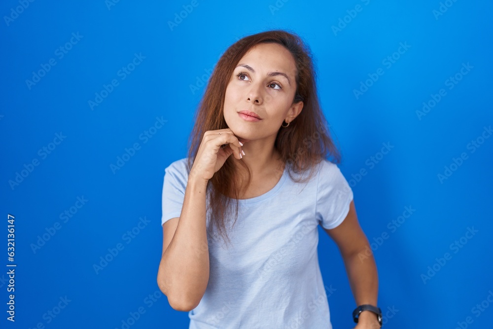 Brunette woman standing over blue background with hand on chin thinking about question, pensive expression. smiling with thoughtful face. doubt concept. Brunette woman standing over blue background with hand on chin thinking about question, pensive expression. smiling with thoughtful face. doubt concept.