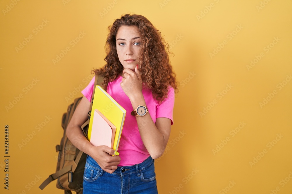 Young caucasian woman wearing student backpack and holding books looking confident at the camera smiling with crossed arms and hand raised on chin. thinking positive. Young caucasian woman wearing student backpack and holding books looking confident at the camera smiling with crossed arms and hand raised on chin. thinking positive.