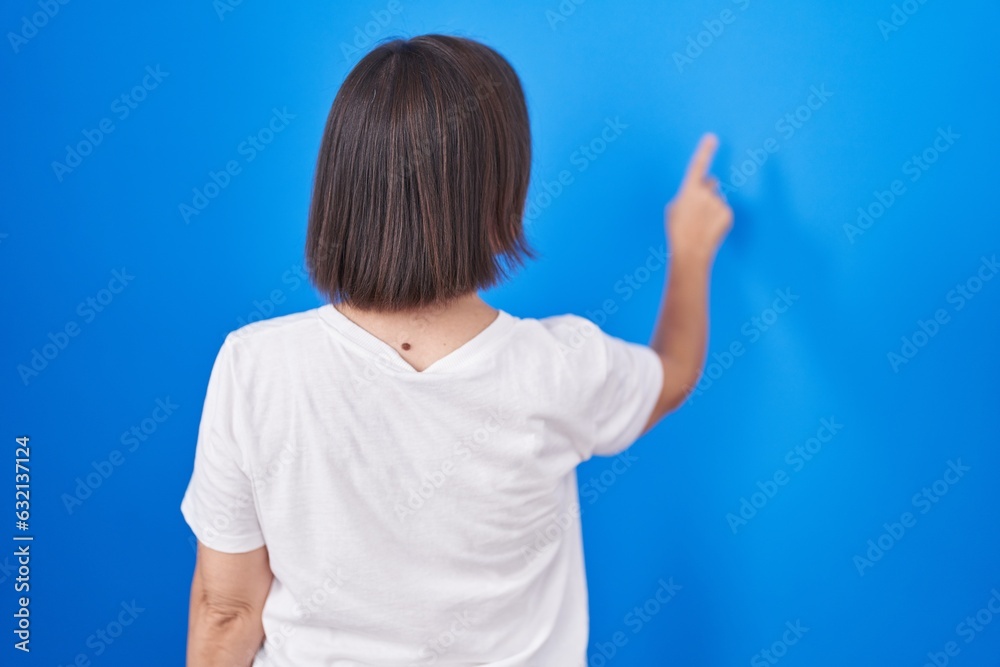 Middle age hispanic woman standing over blue background posing backwards pointing ahead with finger hand Middle age hispanic woman standing over blue background posing backwards pointing ahead with finger hand