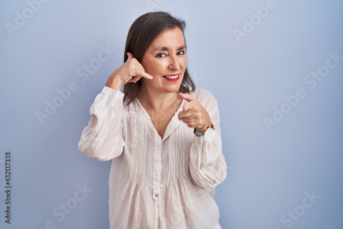 Middle age hispanic woman standing over blue background smiling doing talking on the telephone gesture and pointing to you. call me.