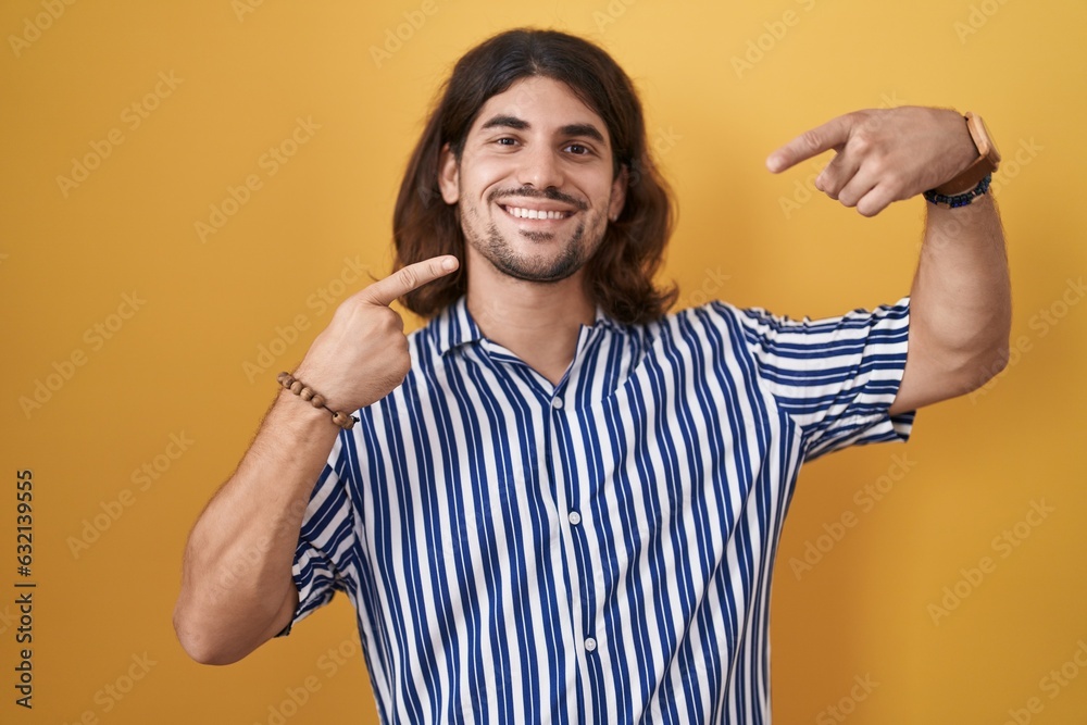 Hispanic man with long hair standing over yellow background smiling cheerful showing and pointing with fingers teeth and mouth. dental health concept. Hispanic man with long hair standing over yellow background smiling cheerful showing and pointing with fingers teeth and mouth. dental health concept.