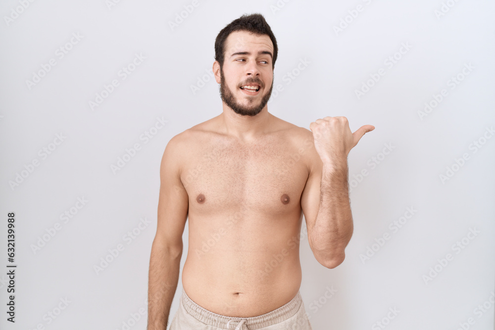 Fototapeta premium Young hispanic man standing shirtless over white background smiling with happy face looking and pointing to the side with thumb up.