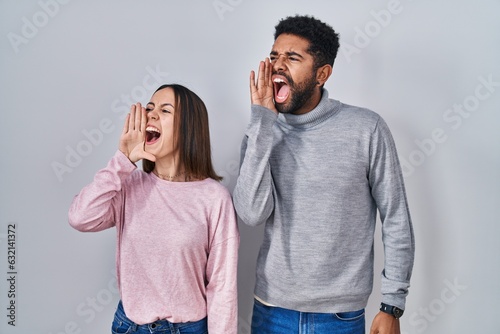 Young hispanic couple standing together shouting and screaming loud to side with hand on mouth. communication concept.